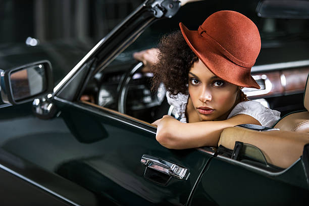 Fashionable retro African American young woman is sitting in an old timer car and looking at the camera.