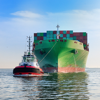 front view on a tugboat towing a large cargo container ship with piled up cargo containers on deck into harbour