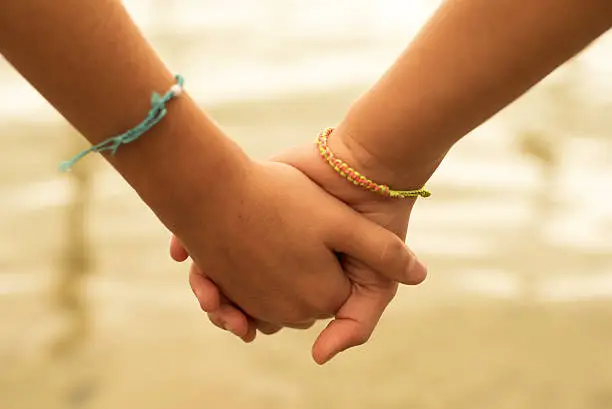 Close up of two children holding hands on the beach sand, summertime friendship concept.