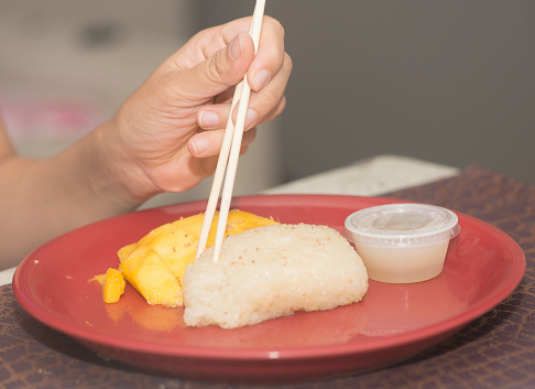 Thai Desert, Sweet sticky rice with fresh mangos and dipping sauce with a female hand holding chopsticks