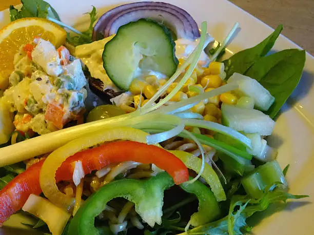 Photo showing a large mixed side salad, consisting of a mixture of vegetables and drizzled in a homemade salad dressing.  Pictured on the plate are chopped pieces of cucumber, potato salad, coleslaw, lettuce, rocket leaves, sliced bell peppers (red, yellow and green), slices of mild red onion, sweetcorn and pasta twirls, with a spring onion garnish on the top of the salad meal.  This salad is part of a healthy eating, low fat diet.