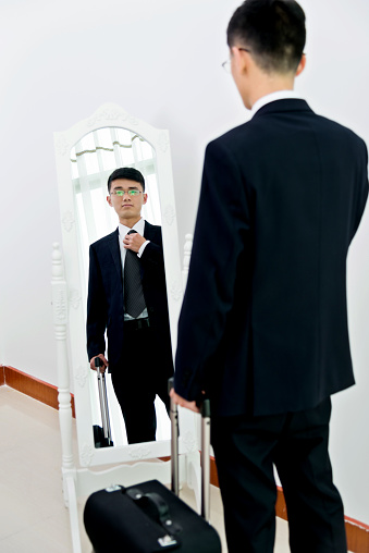 Young asian businessman holding a suitcase and adjusting his necktie in front of mirror.
