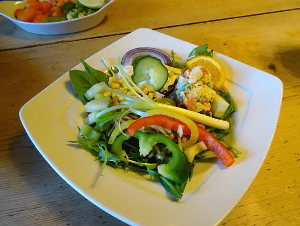 Photo showing a large mixed side salad, consisting of a mixture of vegetables and drizzled in a homemade salad dressing.  Pictured on the plate are chopped pieces of cucumber, potato salad, coleslaw, lettuce, rocket leaves, sliced bell peppers (red, yellow and green), slices of mild red onion, sweetcorn and pasta twirls, with a spring onion garnish on the top of the salad meal.  This salad is part of a healthy eating, low fat diet, being served on a wooden table top.