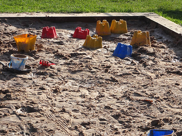 Photo showing a raised sandpit / playpit in a park, with plastic buckets and spades, sandcastles and mounds of sand.  This raised sandpit is edged with old, reclaimed railway sleepers, which contains the fine sand and prevents it from spreading over the lawn grass.