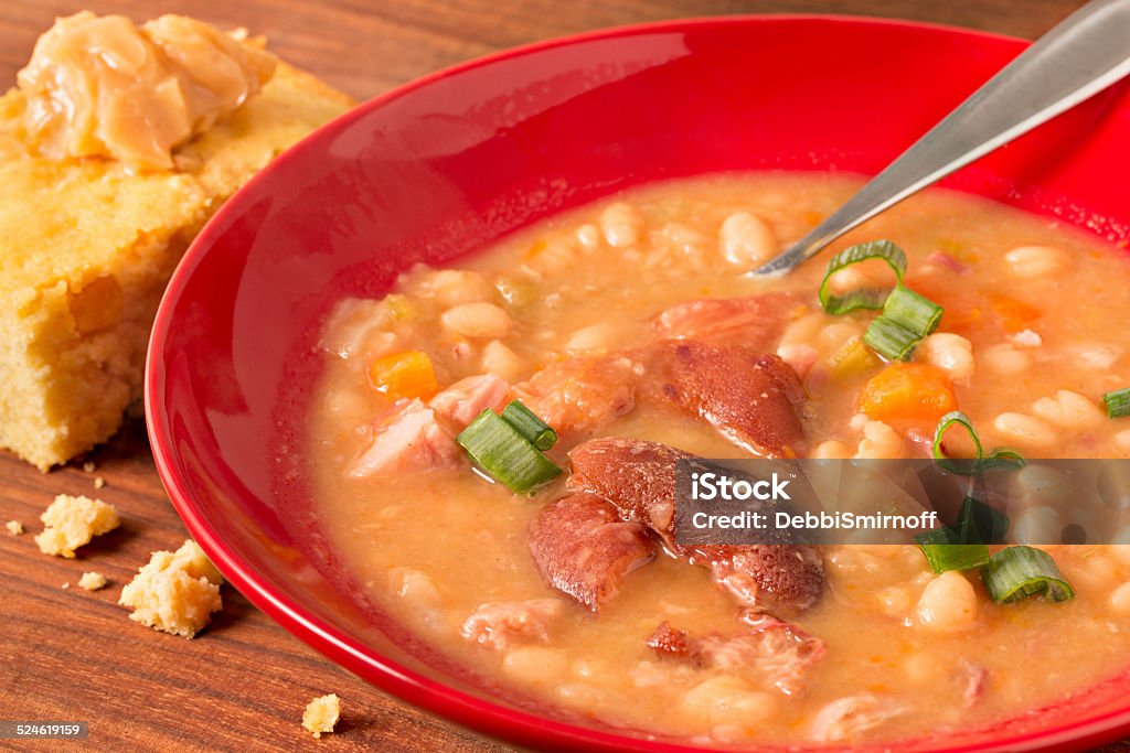 Navy Bean Soup In A Red Bowl A high angle extreme close up shot of a red bowl full of fresh navy bean soup. The soup is choke full of beans, ham hock meat, ham, onions and carrots,  it is garnished with some green scallions. A slice of homemade, freshly baked corn bread with a dollop of honey butter sits by the bowl. Baked Stock Photo