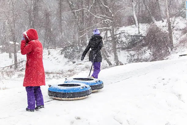 Two sisters have fun with snowtubing at the winter resort. They go to a hill top, oldest sister carry the tube. 