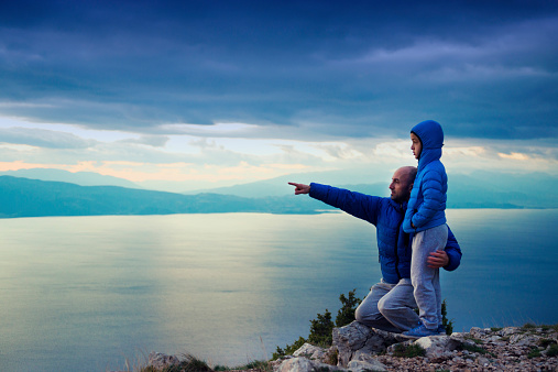 Father And Son On Mountain Hike over the Sea. Pointing to the sunset.