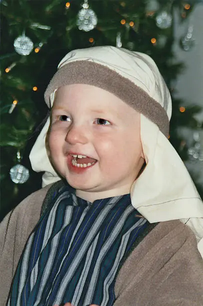 Photo showing a happy young boy dressed as a shepherd, laughing as he performs in his Christmas Nativity play, at nursery school.  A Christmas tree with fairy lights, decorations and silver baubles can be seen in the background.