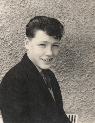 Old black and white photo / retro photograph showing a handsome teenage boy (15 years old) in the late 1950s, posing for the camera with a quiff hairstyle.  His hair has been combed in place with lots of wax / gel / sugar water.  He is pictured smartly dressed, wearing a jacket and tie, just after coming home from school.