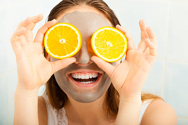 Woman with mud facial mask holds orange slice Beauty skin care cosmetics and health concept. Young woman with facial clay mask holding orange fruit slice covering eyes in bathroom oranges with faces stock pictures, royalty-free photos & images