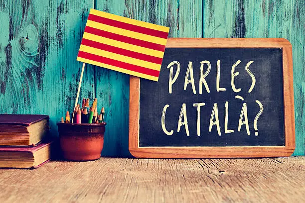 a chalkboard with the question parles catala? do you speak Catalan? written in Catalan, a pot with pencils, some books and the flag of Catalonia, on a wooden desk
