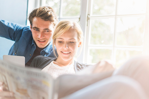 A photo of young couple reading magazine by window. Handsome man with pretty lady spending leisure time at home. Both are in brightly lit area.