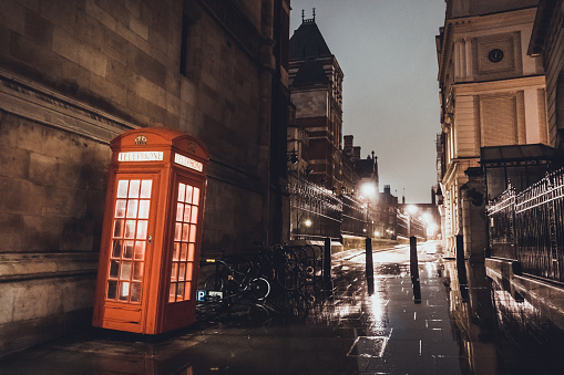 Illuminated red telephone booth on a British city street in a busy commercial district at night on a wet rainy day