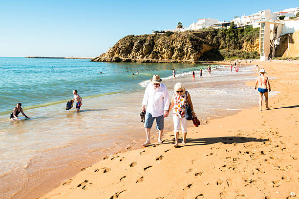 Happy senior couple walking together on a beach Albufeira, Portugal - October 22, 2015: A happy senior couple is walking together on the beach of Albufeira in Portugal while others are (sun)bathing.  albufeira photos stock pictures, royalty-free photos & images