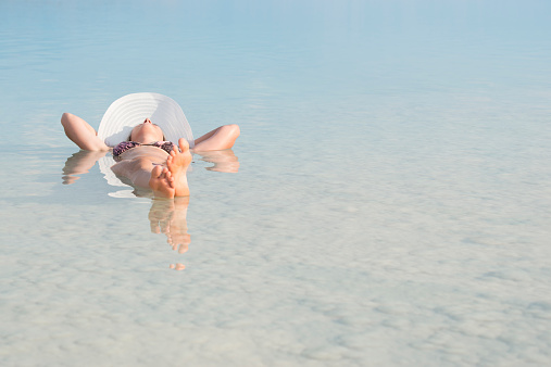 Young woman relaxing, laying on back in salty water with hands behind her head looking up, floating at Dead Sea.