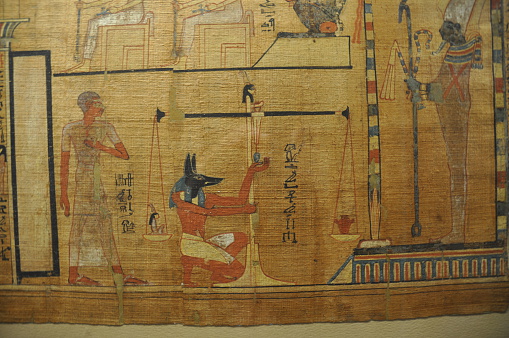 Authentic scene depicted on papyrus showing the climax of the journey to the afterlife. The heart of the deceased is being weighed against Maat, the goddess of justice and truth. On the right, Osiris, god of the underworld and rebirth, presides over the scene. Jackal-headed Anubis, overseer of mummification, adjusts the scales.
