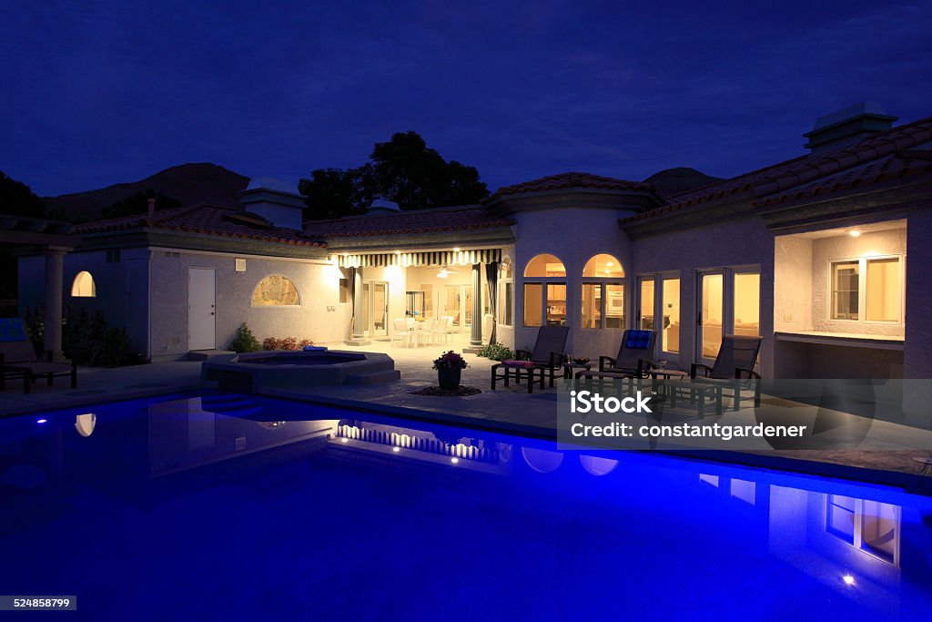 Stunning Pool And Home With Lights At Night 1980`s Spanish Mediterranean  architecture. Evening on the luxurious back pool and patio. Lights in home and outside. Lounge chairs poolside. Dining area in covered patio. Night Stock Photo