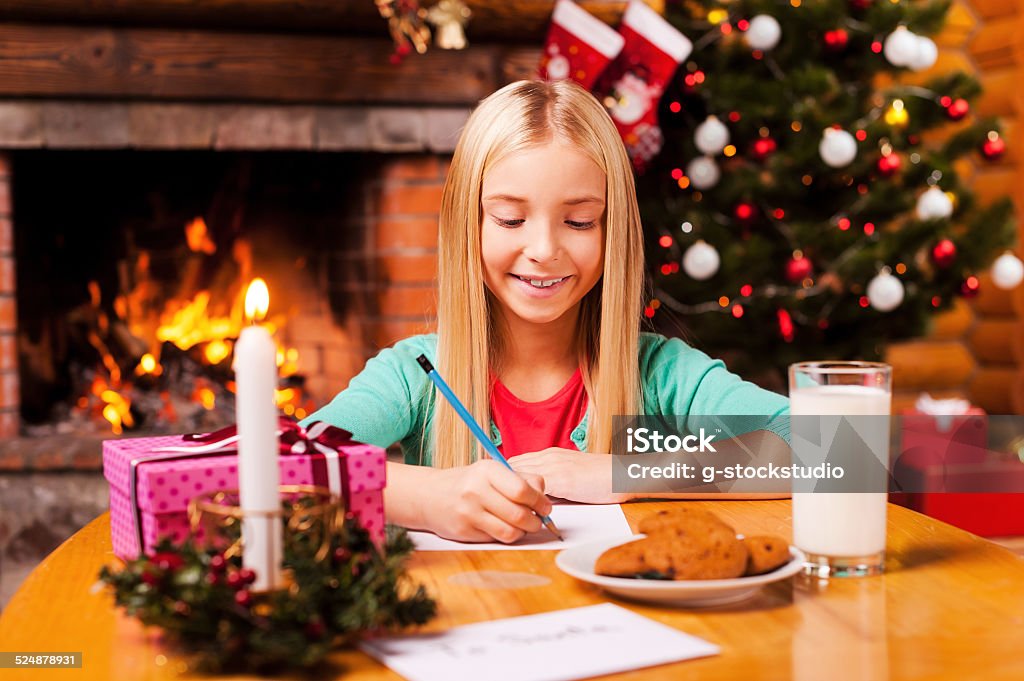 Writing a letter to Santa. Cute little girl writing a letter to Santa Claus while sitting at home with Christmas tree and fireplace in the background 6-7 Years Stock Photo