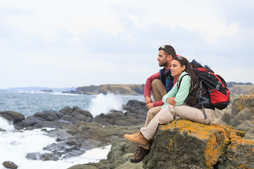 Young couple sitting on rock and resting after long hiking. Looking at  waves crashing into the rocks. Both with sunglasses, backpacks, hiking boots and casual clothes.