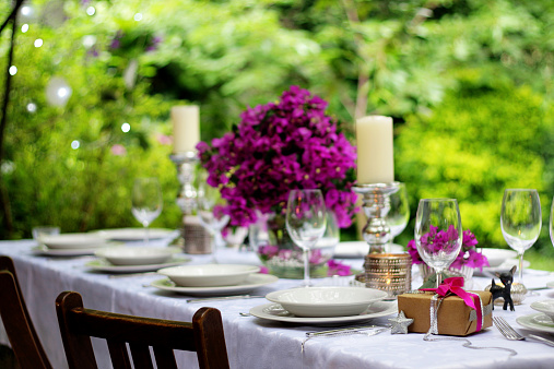 A DSLR photo of an elegant table setting for an outdoor summer dinner party at the garden during afternoon. Lush green foliage and Christmas lights defocused at the background. Bougainvillea flower arrangement, candles and silver decoration. White tableware. White tablecloth. Wooden chairs. Wine glasses. Beautiful gift box at the foreground on the table. Main colours are green, silver, white and pink. Christmas celebration in southern hemisphere countries.