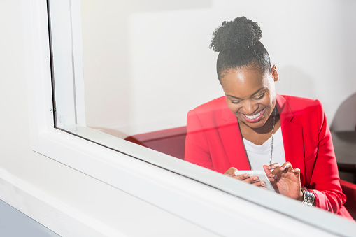 A mid adult, stylishly dress black woman using a mobile phone, texting. She is wearing a red jacket and white shirt, hair up in a bun, smiling as she looks down at her smartphone. The view is from the outside looking through a window. She is in a business office.