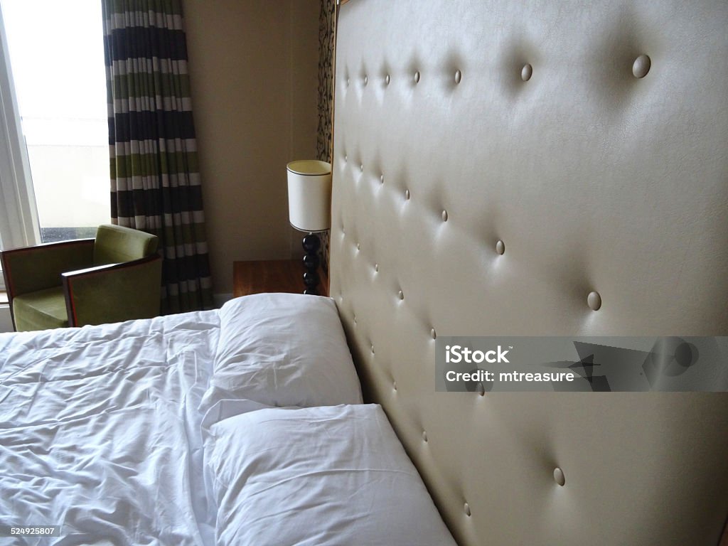 Kingsize double bed, large beige studded leather headboard, white-sheets, bedside-lamp Photo showing a king-size double bed with a large beige studded leather headboard (buttoned / buttons), white duvet cover and sheets, and a wooden bedside lamp on a side table for reading in bed.  Patterns ivory cream and black wallpaper on a feature wall behind the bed adds a luxurious feel to the bedroom suite, blending in with the outsized headboard. Armchair Stock Photo