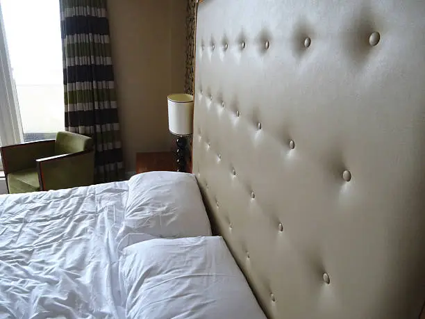 Photo showing a king-size double bed with a large beige studded leather headboard (buttoned / buttons), white duvet cover and sheets, and a wooden bedside lamp on a side table for reading in bed.  Patterns ivory cream and black wallpaper on a feature wall behind the bed adds a luxurious feel to the bedroom suite, blending in with the outsized headboard.