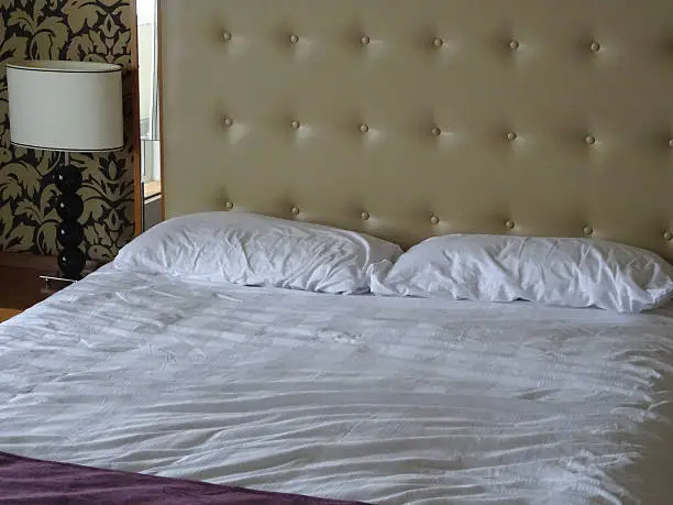 Photo showing a king-size double bed with a large beige studded leather headboard (buttoned / buttons), white duvet cover and sheets, and a wooden bedside lamp on a side table for reading in bed.  Patterns ivory cream and black wallpaper on a feature wall behind the bed adds a luxurious feel to the bedroom suite, blending in with the outsized headboard.