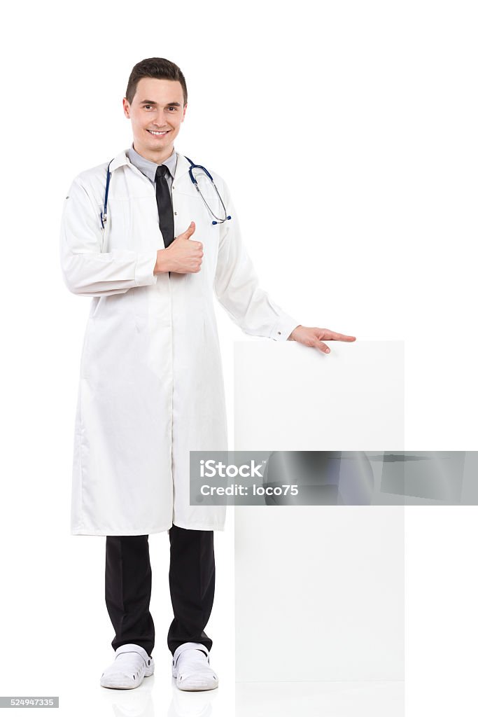 Good medicine Smiling male doctor standing close to white box and showing thumb up. Full length studio shot isolated on white. Doctor Stock Photo
