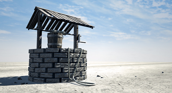 A brick water well with a wooden roof and bucket attached to a rope in a flat barren landscape with a blue sky background