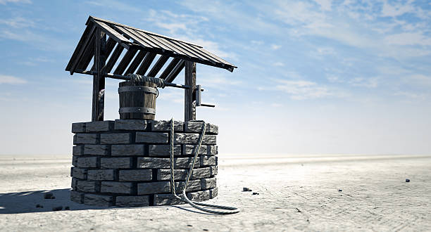 Wishing Well With Wooden Bucket On A Barren Landscape A brick water well with a wooden roof and bucket attached to a rope in a flat barren landscape with a blue sky background wells stock pictures, royalty-free photos & images