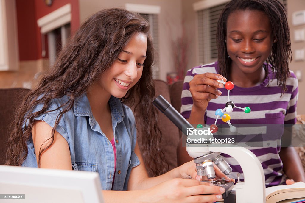 Education.  Teenage girls studying science at home. Homework. Latin and African descent teenage girl friends studying DNA molecular structure, using microscope as they study science subject at home setting.  The multi-ethnic friends hold the model and look into the microscope as they learn, study their science homework together.  They are collaborating on a science project. Open textbook in girls lap.  Kitchen background.  Education themes.  Girls in STEM school classes. 14-15 Years Stock Photo