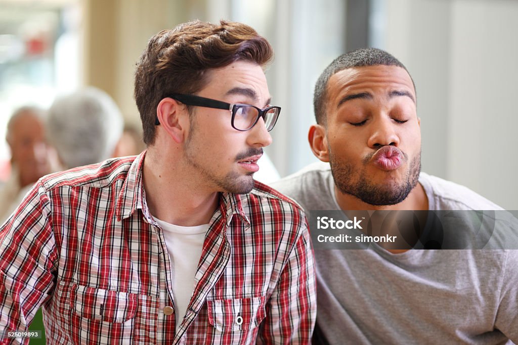 Man Making Funny Faces Young man making funny faces while his friend looking him weird at the table in the cafe.   Gay Man Stock Photo