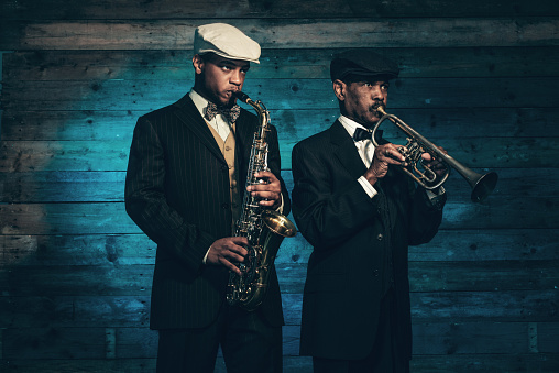 Two vintage african american jazz musicians with trumpet and saxophone in front of old wooden wall. Wearing black suit and cap.
