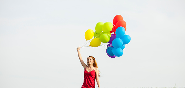 Cheerful young woman with red dress try to hold a brunch of multi colored balloons blown away by the wind.