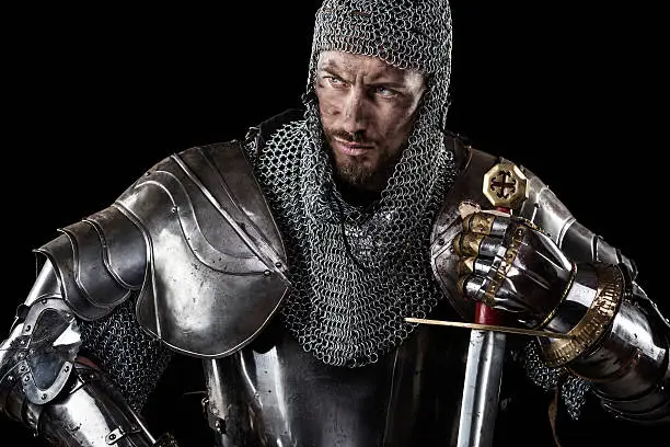 Portrait of Medieval Dirty Face Warrior with chain mail armour and red cross on sword. Dark Background