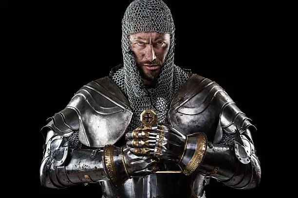 Portrait of Medieval Dirty Face Warrior with chain mail armour and red cross on sword. Black Background