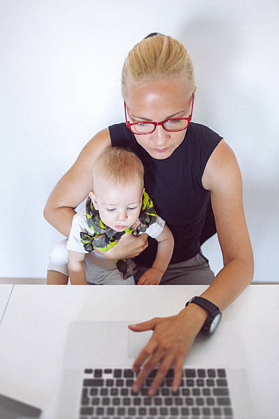 Mother holding her baby while working on laptop from home stock photo