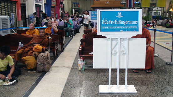 Bangkok, Thailand - March 5, 2016: Looking At Buddhist Monks Sitting Waiting For Train At A Place Where There Is A Sign Board Saying Reserved For Monks And Novices Inside Bangkok's Train Station In Thailand Southeast Asia.Including Lots Of People Sitting Behind Them Also Waiting For Train.In Between The Waiting Time Some Of Them Decided To Eat Food,Make Telephone Call While Some Are Walking And Looking Around
