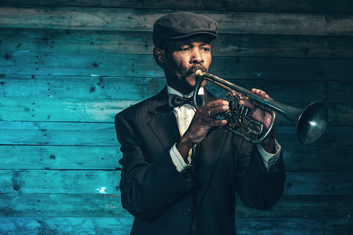 Vintage african american senior jazz musician with trumpet in front of old wooden wall. Wearing black suit and cap.