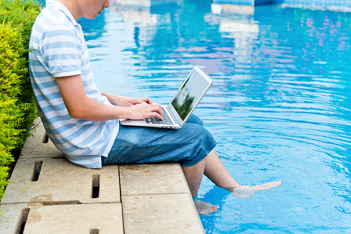 young man working by the pool with feet in the swimming pool