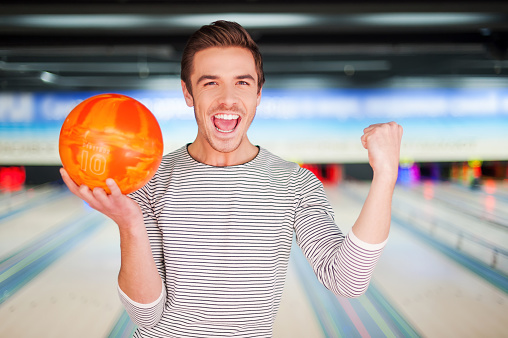 Cheerful young man holding a bowling ball and keeping arms raised while standing against bowling alleys