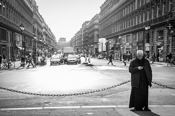 Paris, France - January 2, 2012: A Priest uses his mobile phone in the center of busy street of Place de l'Opéra, Paris, France. People can be seen crossing the road at zebra crossing where traffic stops at red light. In the distance is the Louvre Museum