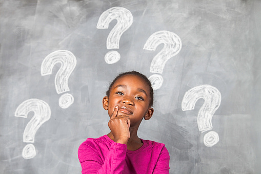 African girl sitting in front of a black chalk board with question marks drawn on it. She it thinking very hard and she has her finger by her mouth. Cape Town, Western Cape, South Africa