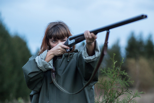 A red haired girl in a raincoat standing outdoors with a shotgun aiming at a target.