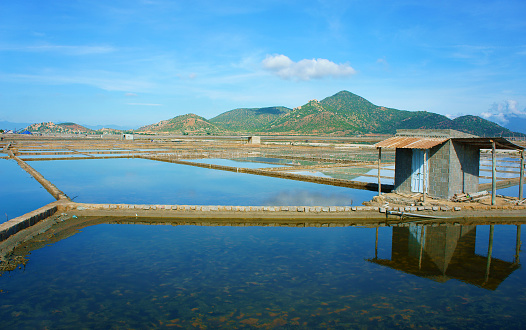 Beautiful landscape of saline filed on day under blue sky, store house reflect on saltwater, chain of mountain behind, amazing scene of agriculture at Vietnamese countryside