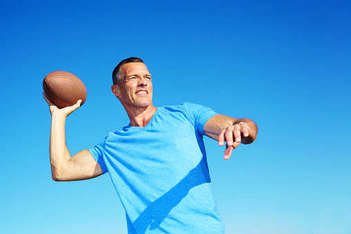 Confident mature man throwing American football against clear blue sky