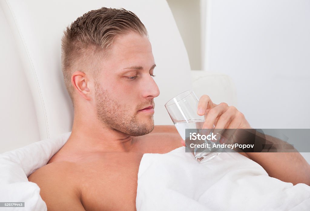 Man drinking a glass of water in bed Man lying in bed propped up against the pillows drinking a glass of fresh water Adult Stock Photo