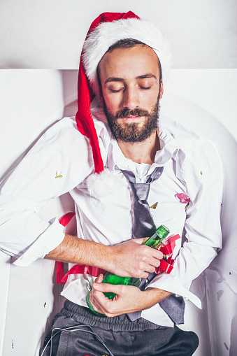 The host of the Christmas party sleeps peacefully in empty bathtub, covered in confetti and holding an empty beer bottle. Christmas party is over.