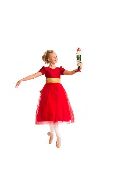 Beautiful dainty Caucasian girl with blond curls wearing a red party dress portrays Clara from the Christmas Nutcracker ballet as she stands en pointe holding a nutcracker doll against a white background